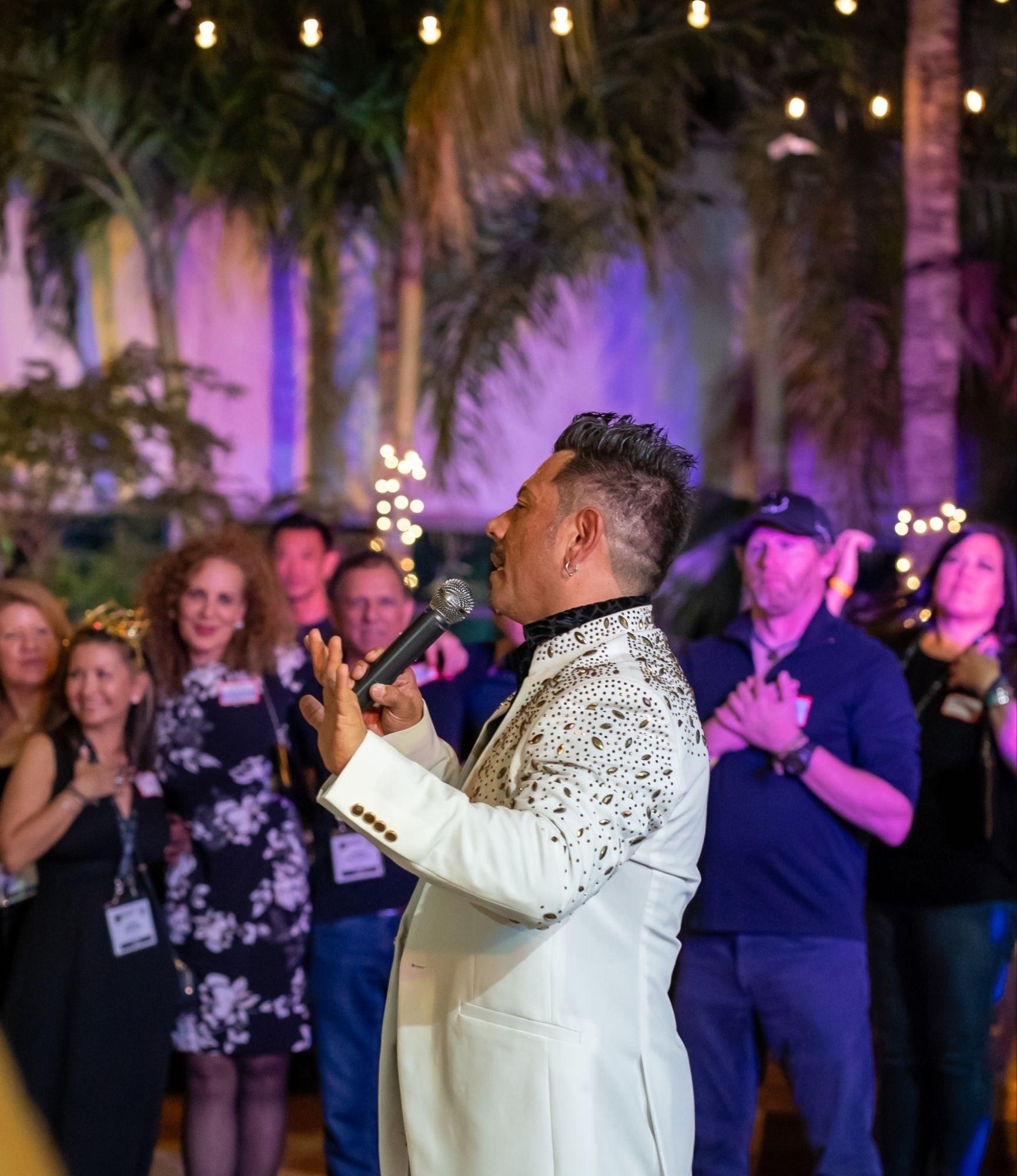J.J. Villar in a white jeweled jacket speaking into a microphone to an evening audience