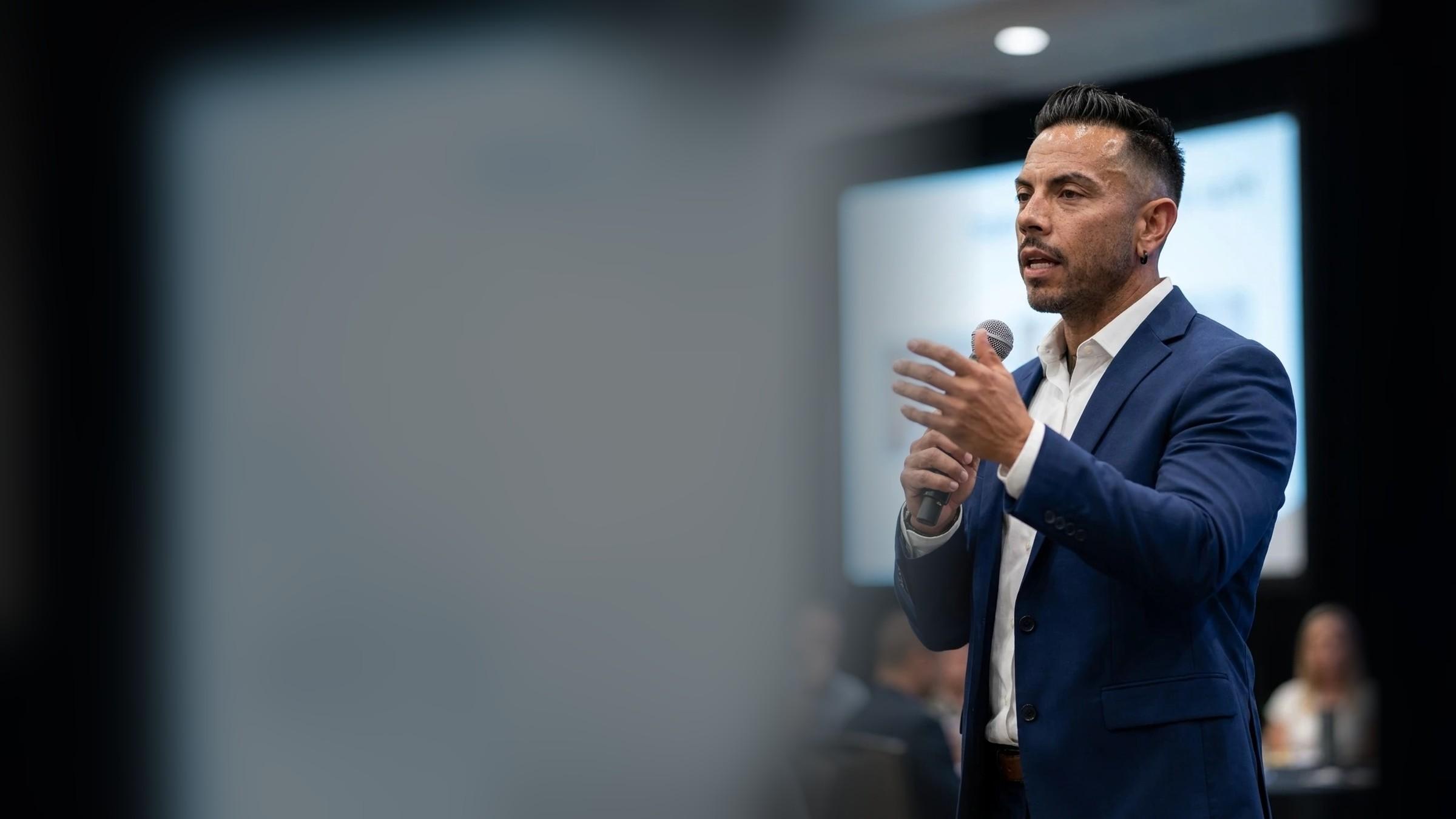 J.J. Villar in a navy blazer speaking into a handheld microphone in a conference room