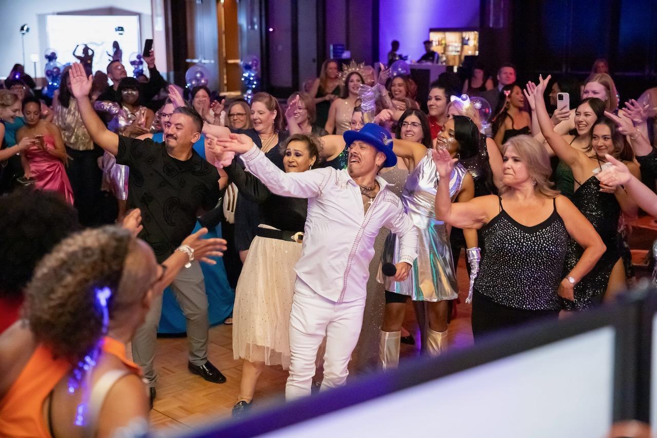 J.J. Villar in a white suit and blue hat leading a ballroom full of guests through choreography