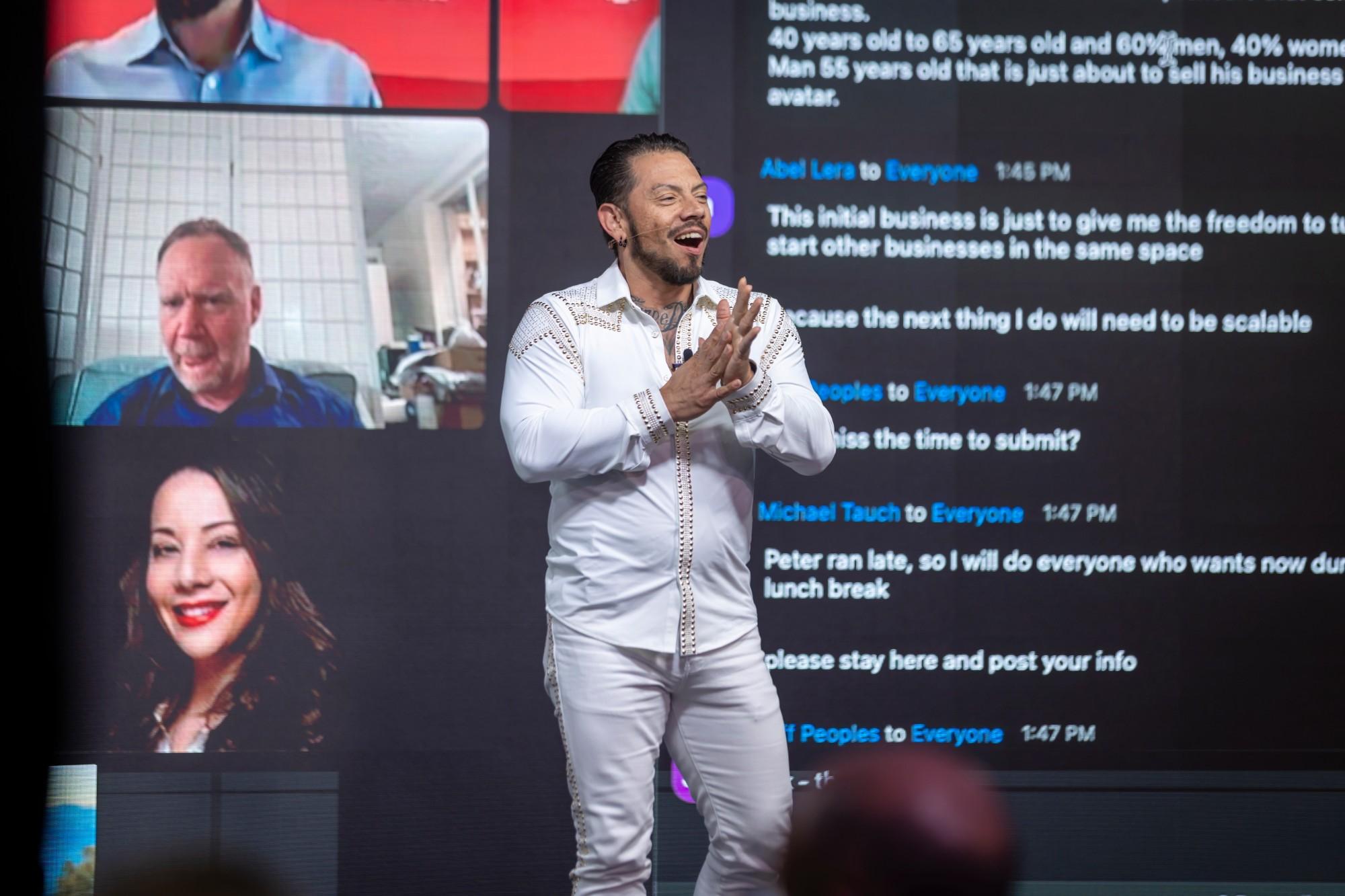 J.J. Villar on stage in a white suit, smiling and clapping with the room