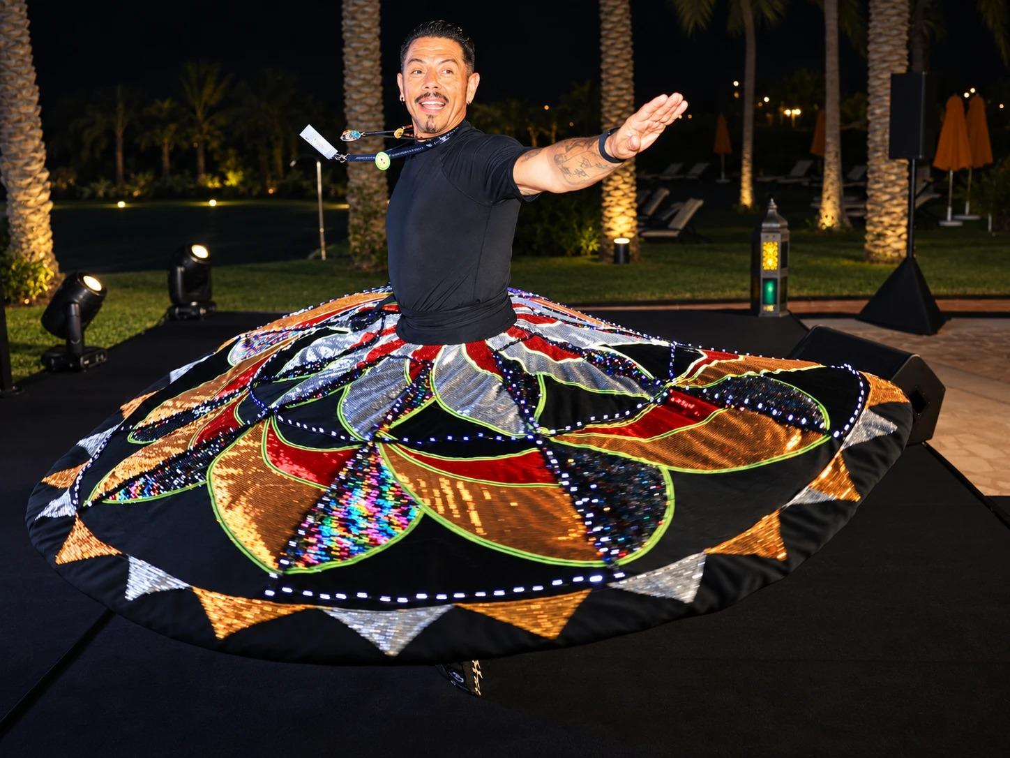 J.J. Villar mid-spin in a lit, sequined tanoura skirt, smiling and holding his focus