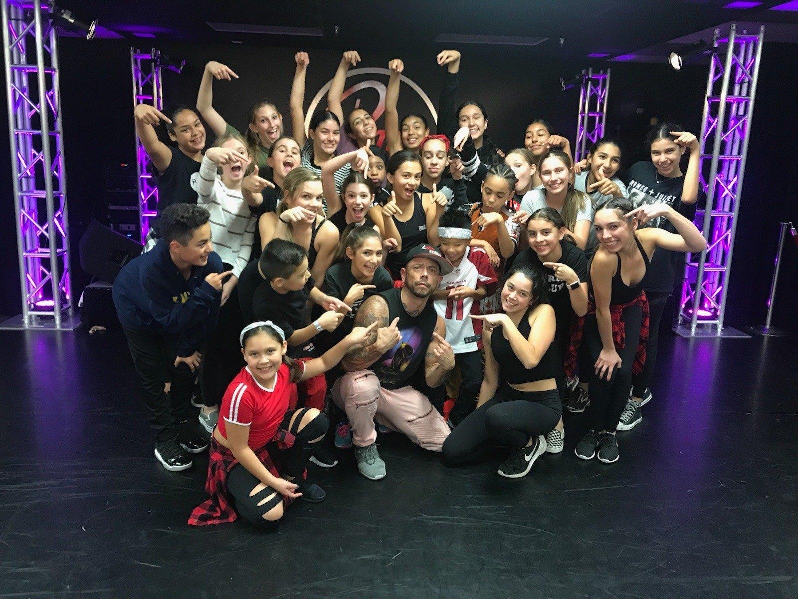 J.J. Villar crouched center stage with a large group of young dancers all pointing at him